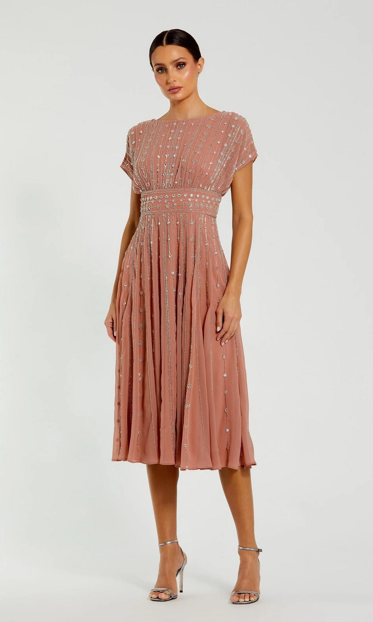 A woman stands in a cinnamon chiffon cocktail dress, featuring short sleeves and delicate beadwork. The Midi Party Dress: Mac Duggal 94153 showcases graceful pleats and pairs beautifully with her silver high-heeled sandals, all set against a plain background.